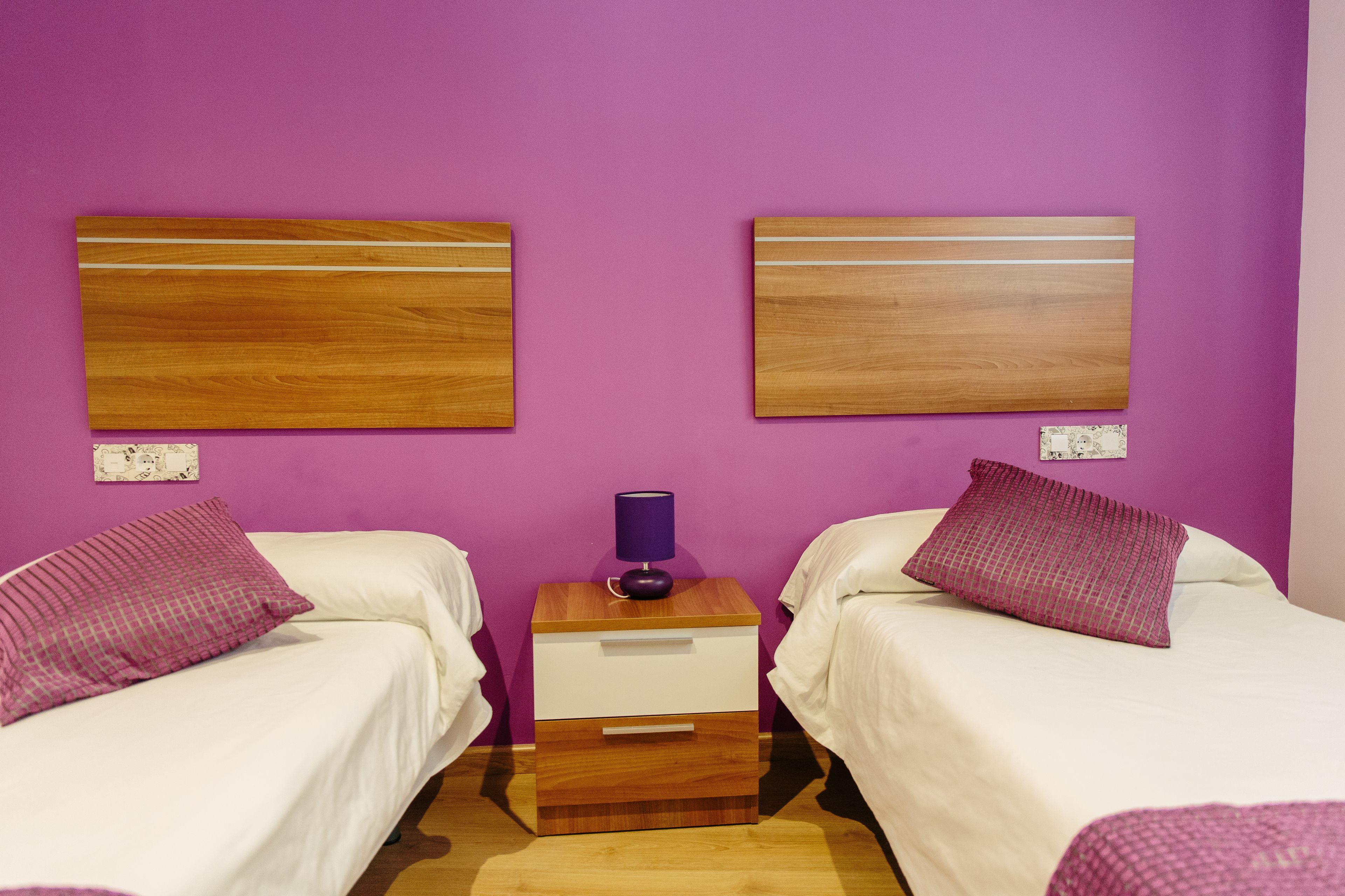 A room with two beds, each with a wooden headboard, a nightstand with a lamp, and a small drawer.