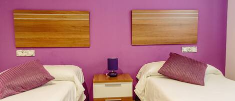 A room with two beds, each with a wooden headboard, a nightstand with a lamp, and a small drawer.