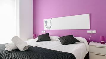 A modern bedroom with a purple wall, a large bed with white and black bedding, two bedside tables with lamps, and a wall-mounted light fixture.