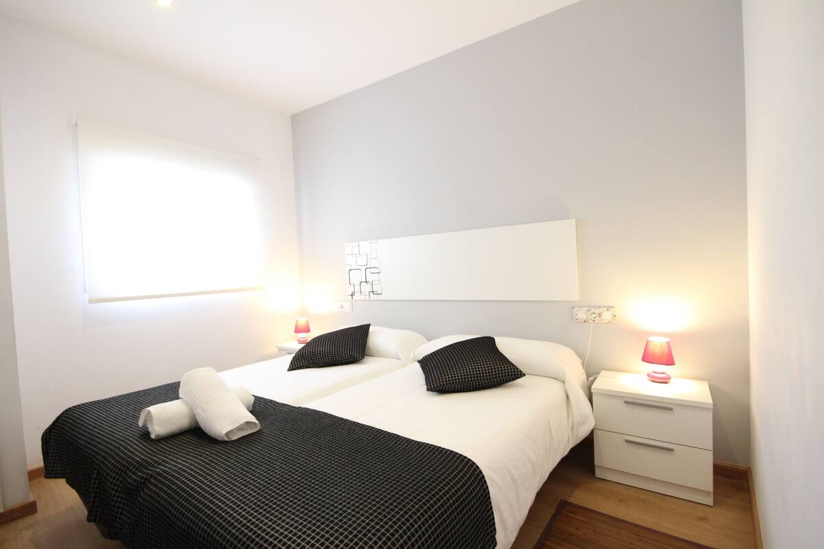 Een hotelkamer met twee bedden, een nachtkastje met een lamp en een raam met jaloezieën.