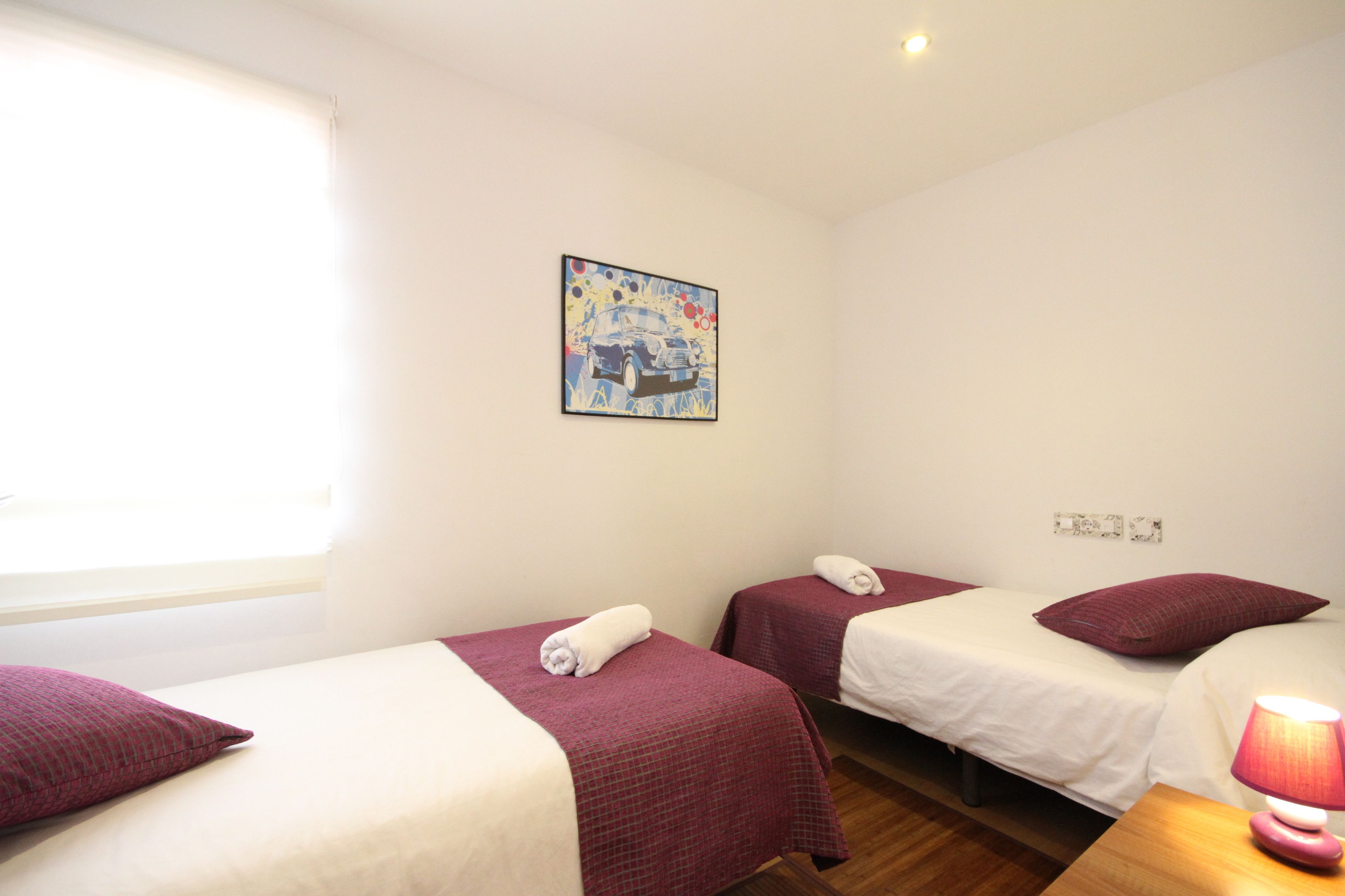 A hotel room with two single beds, each with a red bedspread and white linens, a small table with a lamp, and a painting on the wall.
