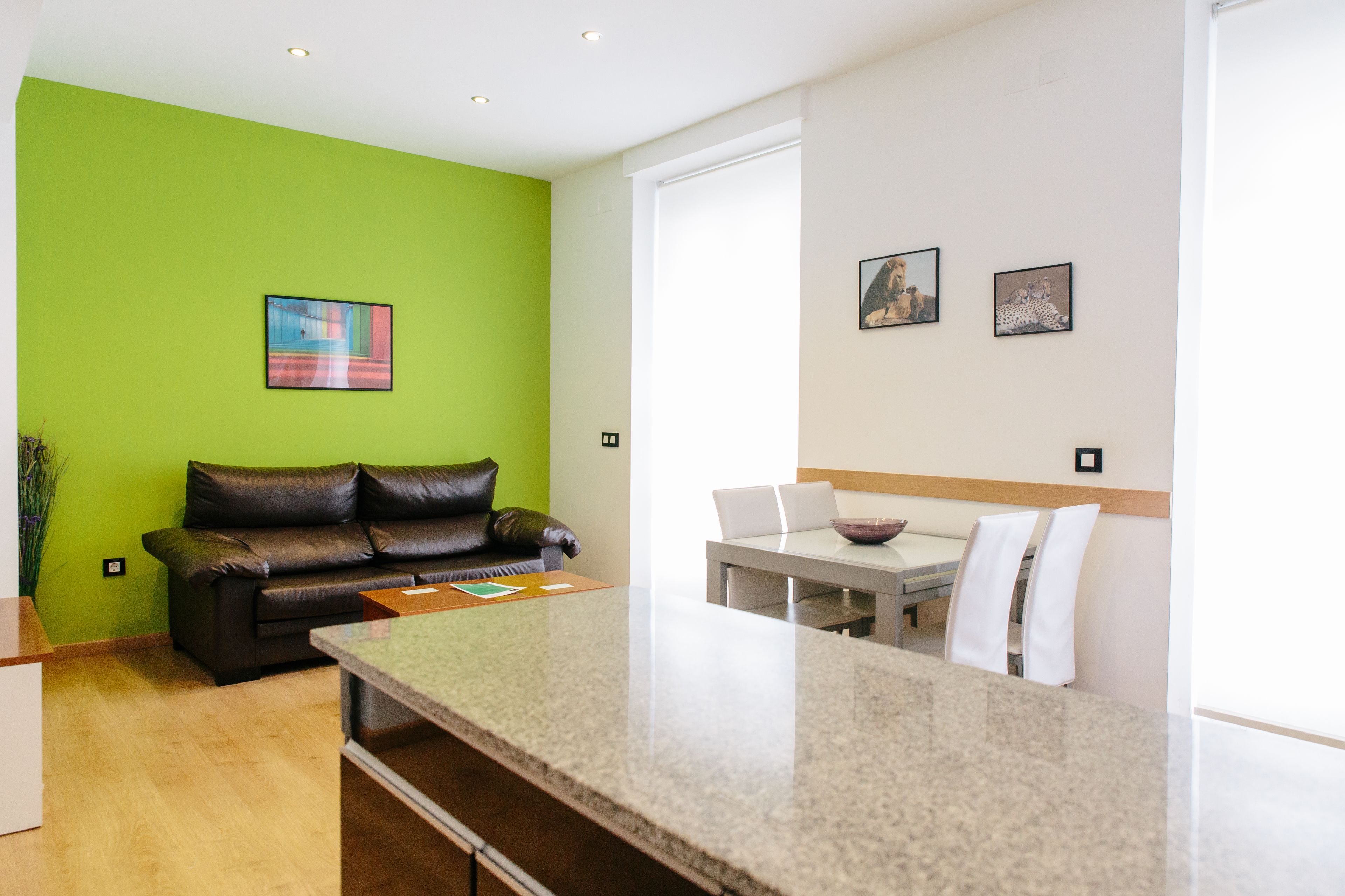 Una sala moderna con una pared verde de acento, un sofá de cuero marrón, una zona de comedor con una mesa y sillas blancas, y una cocina con encimera de granito.
