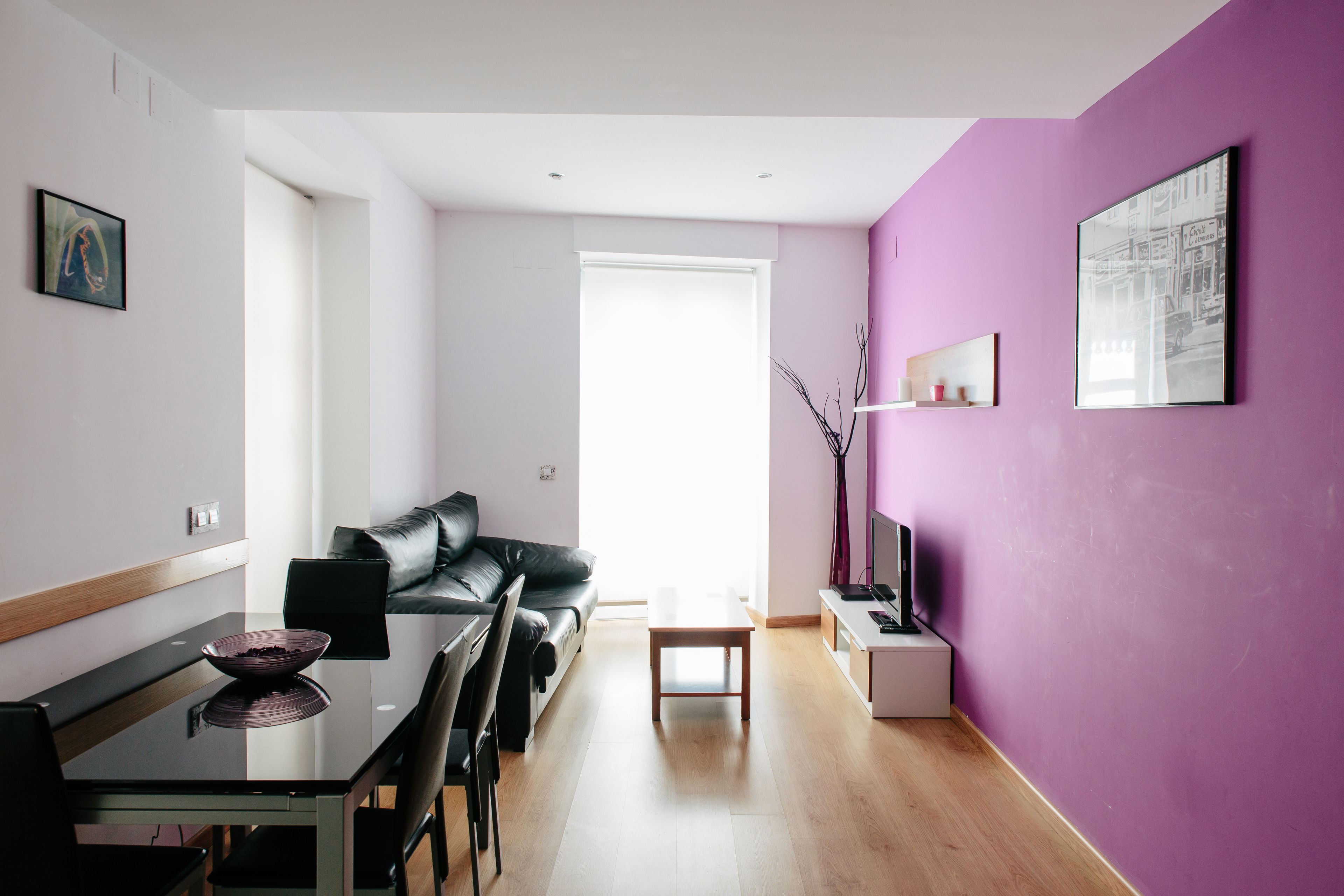 A modern living space with a dining area, a black leather sofa, a glass table, and a purple accent wall.