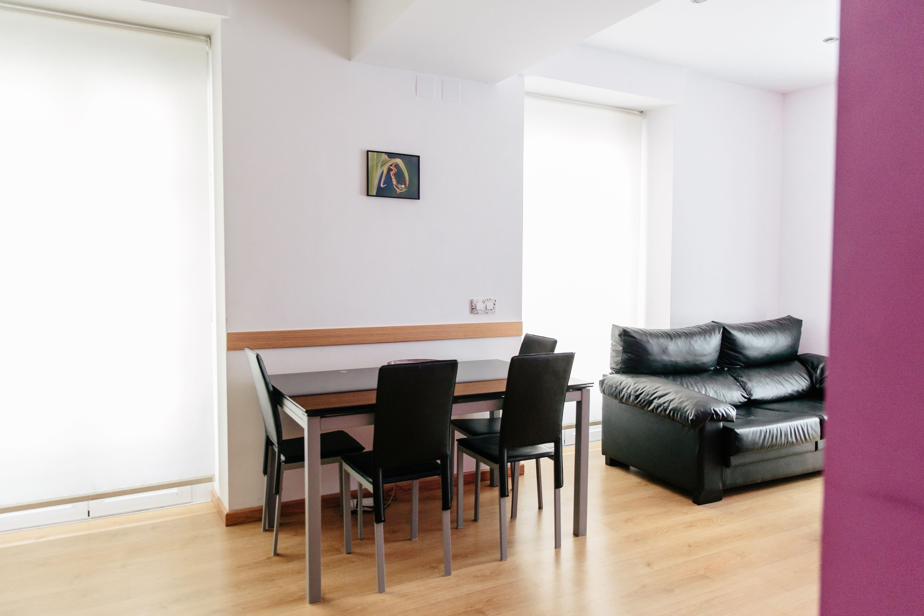 A modern living room with a dining area, featuring a wooden dining table, black chairs, a black leather sofa, and a framed artwork on the wall.