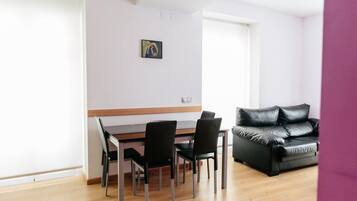 A modern living room with a dining area, featuring a wooden dining table, black chairs, a black leather sofa, and a framed artwork on the wall.