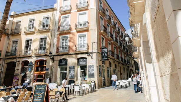 A café with outdoor seating, a palm tree, and a building with balconies.