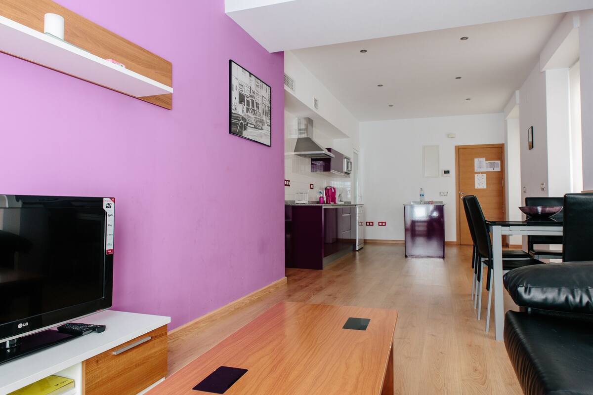 Een moderne woonkamer met een flatscreen televisie, houten vloeren, een eethoek met een tafel en stoelen, en een keuken op de achtergrond.