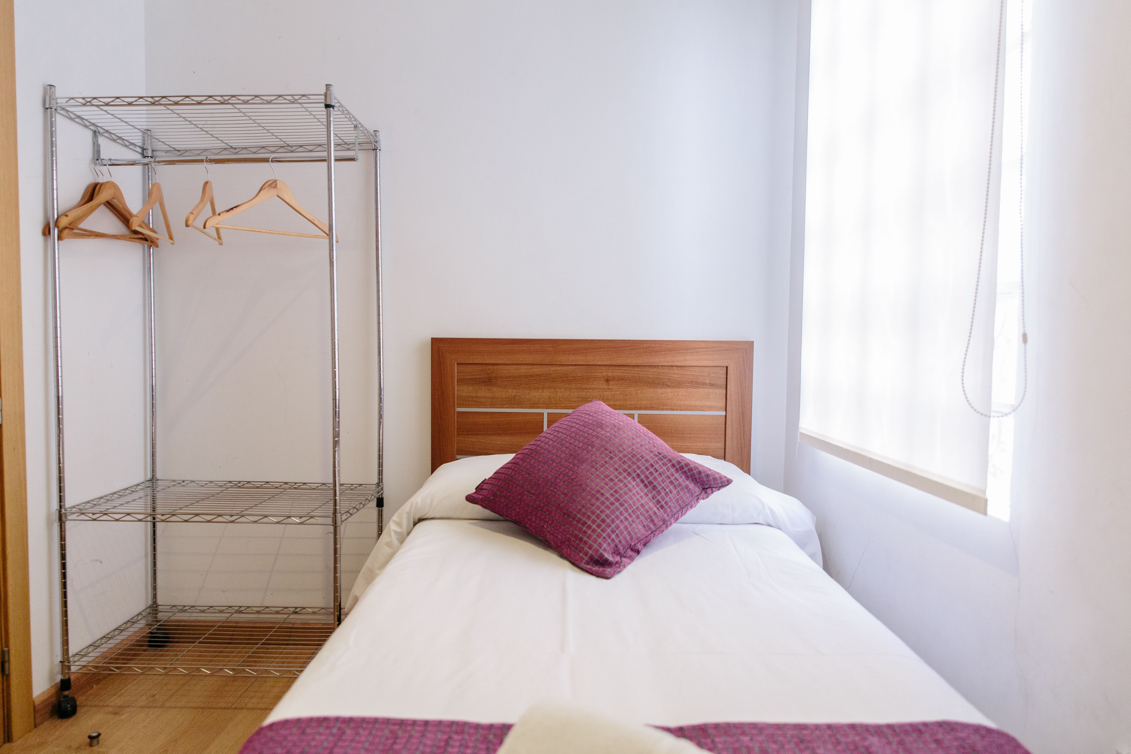 A single bed with a wooden headboard, a purple pillow, and a metal shelf unit with wooden hangers.