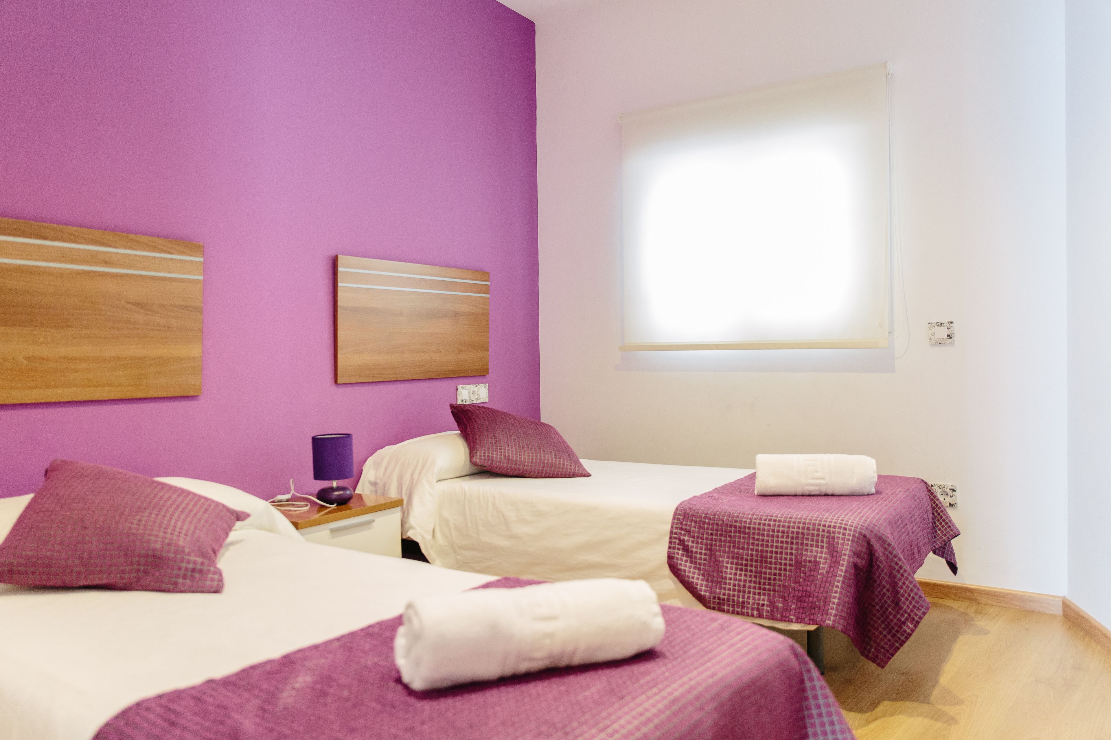 A hotel room with two beds, purple bedding, a small table with a lamp, and a window with blinds.