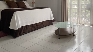 Un dormitorio con cama, una mesita de vidrio, una cortina y ventana con vistas a follaje.