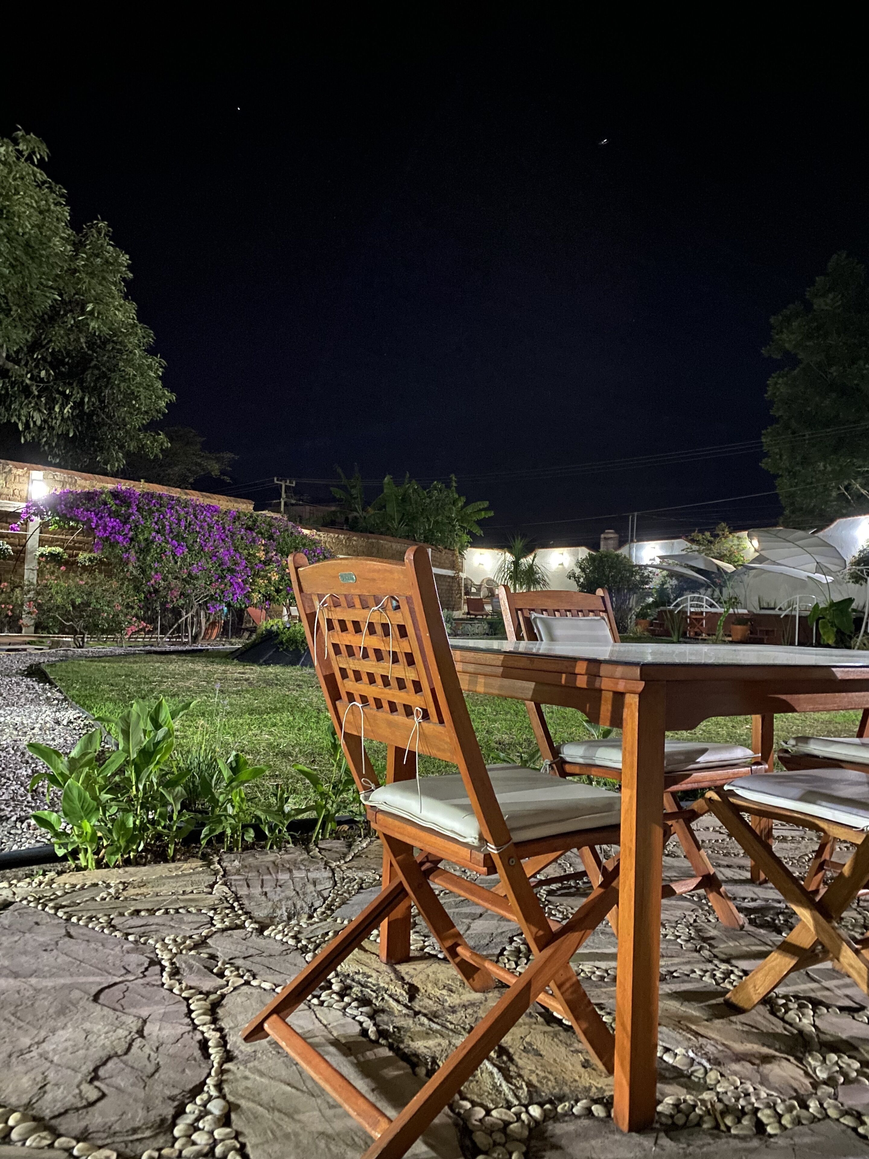 An outdoor dining area with wooden chairs and a table, surrounded by a garden with plants and a stone pathway.