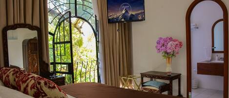 A hotel room with a bed, a TV, a window with curtains, a vase with flowers, and a mirror.