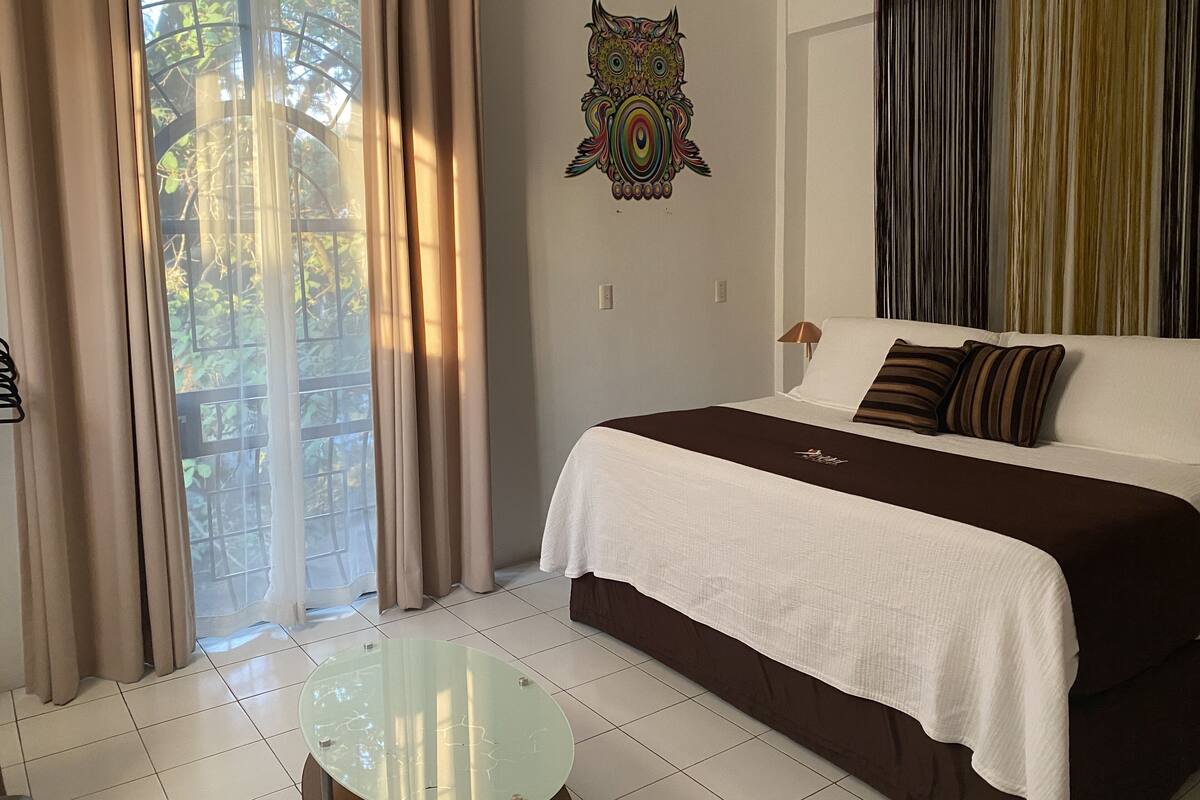 Una habitación de hotel con cama, una mesita de vidrio, cortinas y un adorno en la pared.