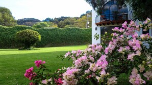 Un jardín exuberante con flores vibrantes rosa y blanco en primer plano, un césped cuidado y un edificio blanco con balcón al fondo.