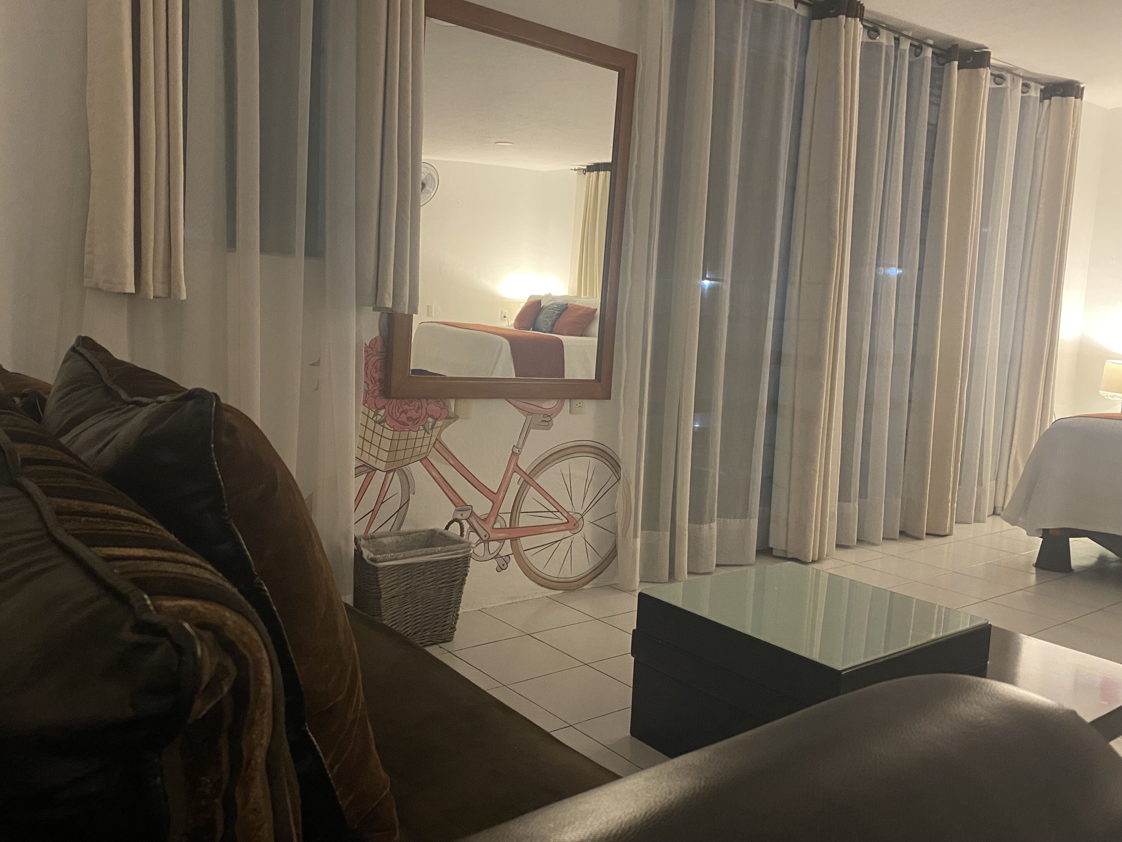 A hotel room with a large mirror, a bicycle-themed wall decoration, a sofa, a small table, and a lamp.
