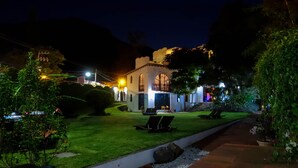 Un edificio bien iluminado con jardín, mesas al aire libre y una escalera que conduce a la entrada.