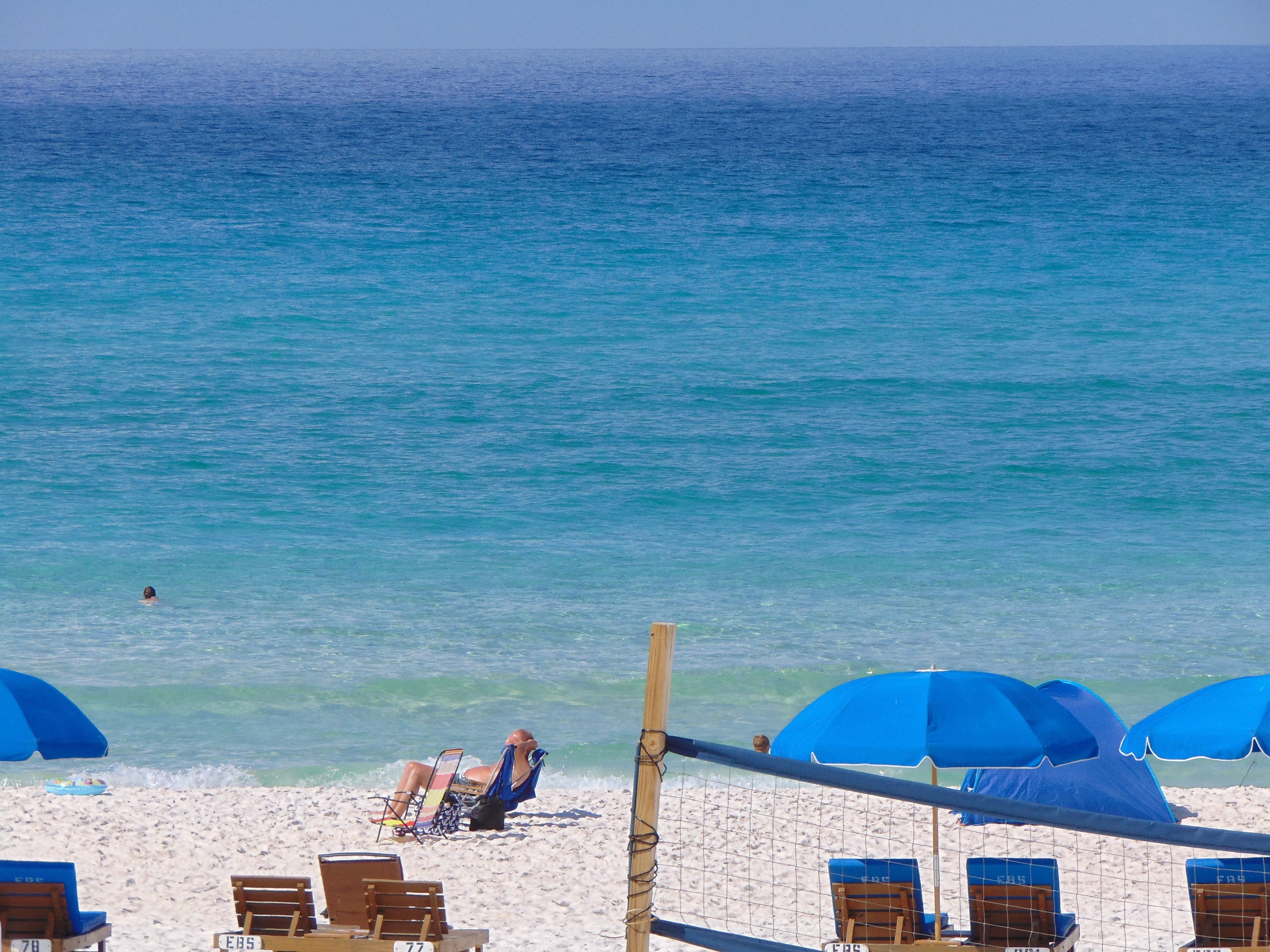 A beach with a volleyball net, blue umbrellas, and beach chairs.