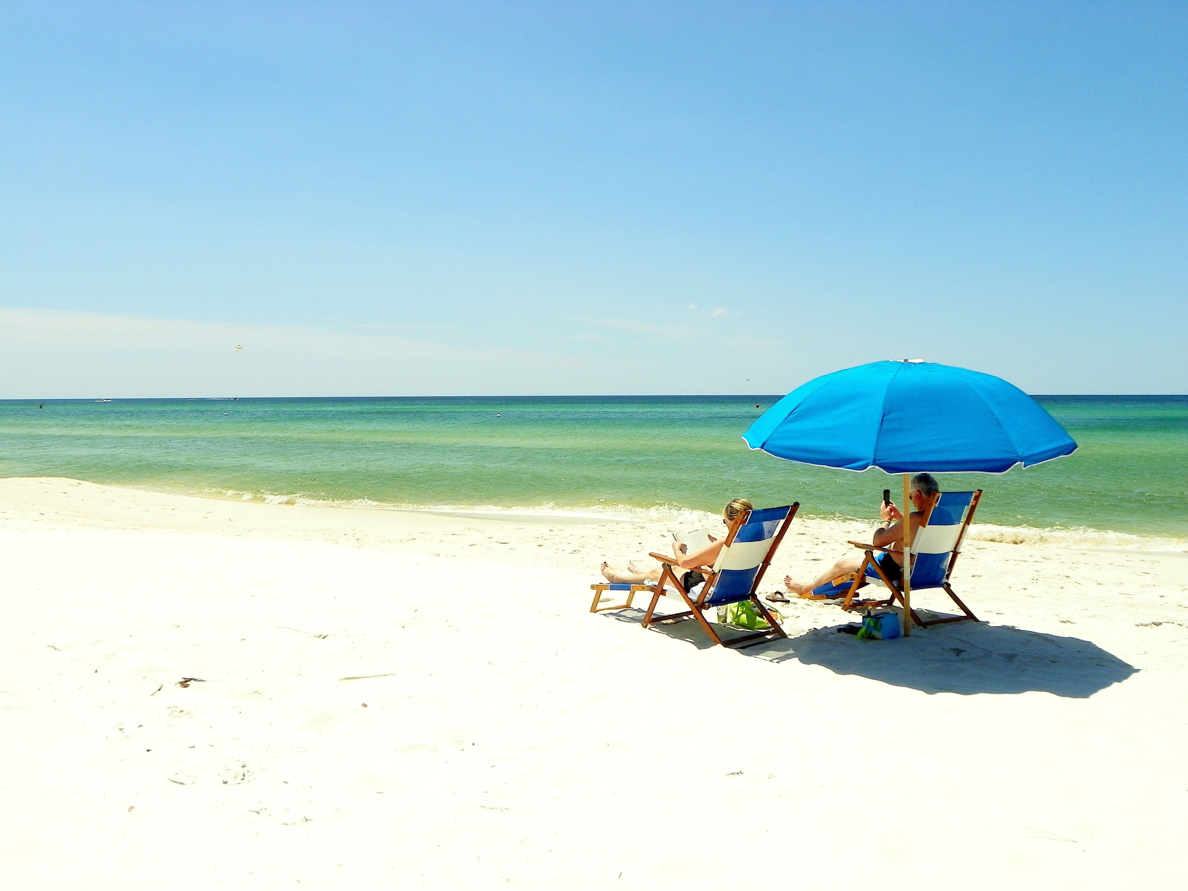 Zwei Strandstühle unter einem blauen Sonnenschirm an einem sandigen Strand mit klarem Wasser.