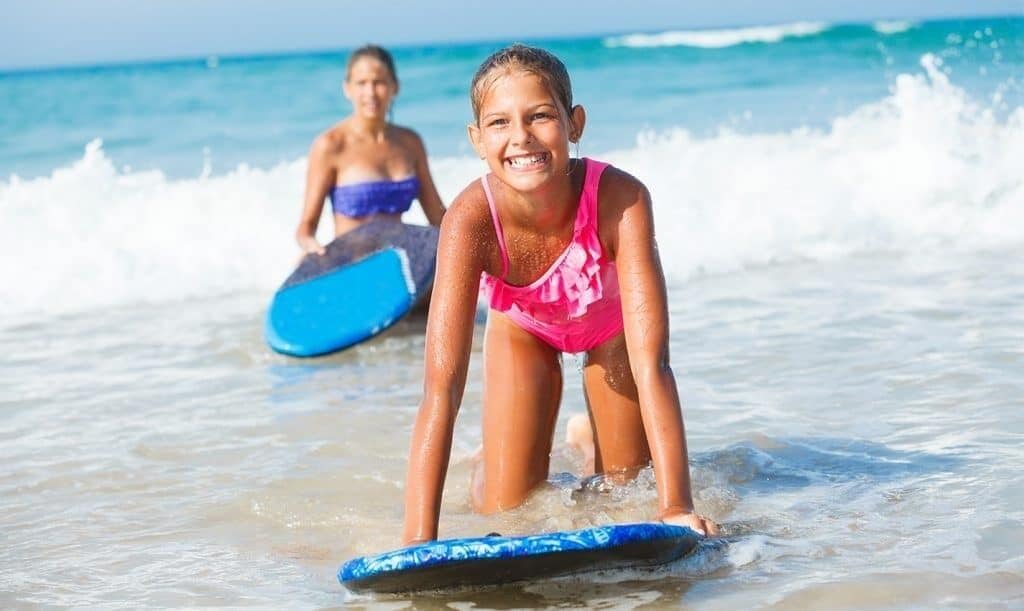Une personne portant un maillot de bain rose, debout sur une planche de surf bleue, dans l’eau.