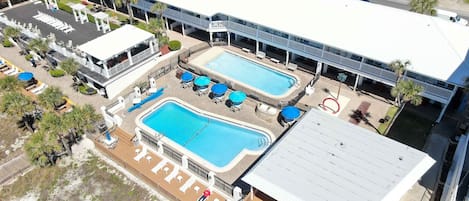 An aerial view of a hotel with two swimming pools, a covered deck area, and a parking lot.
