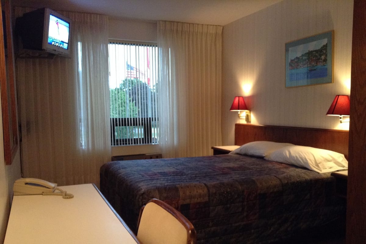 A hotel room with a bed, a desk with a telephone, two bedside lamps, a window with curtains, and a television mounted on the wall.