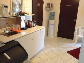 A small kitchenette with a microwave, sink, and storage cabinets. There is a water dispenser and a chair in the corner.