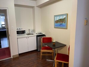 A compact kitchen with white cabinets, a microwave, and a sink. There is a dining area with a small table and two chairs. A painting of a coastal scene adorns the wall. - Shakespeare Inn (Perth East)