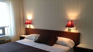 Una habitación de hotel con una cama grande, dos lámparas de noche y un ventanal con cortinas.