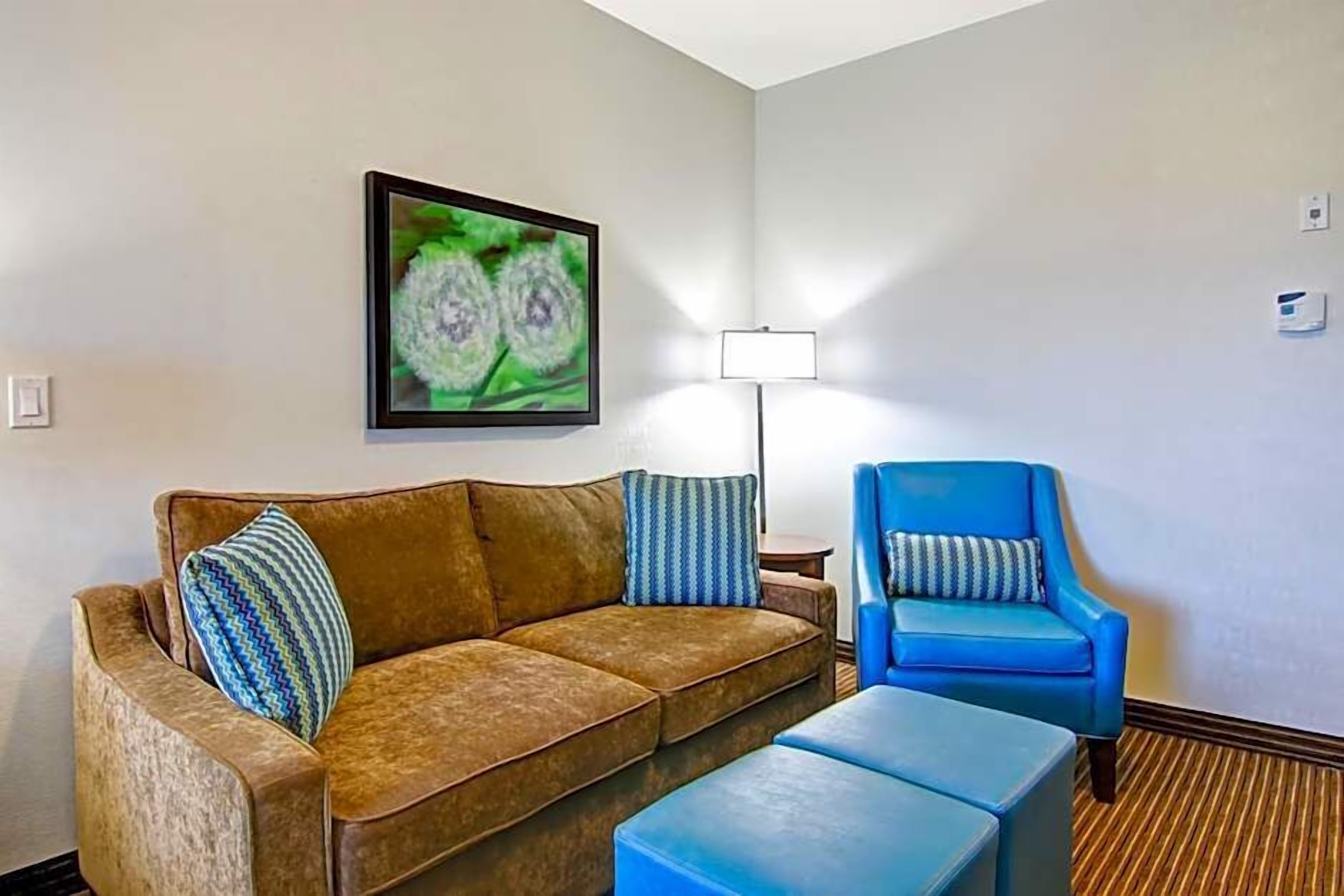 A living room with a brown sofa, a blue armchair, and a small table. There is a framed picture on the wall and a floor lamp.