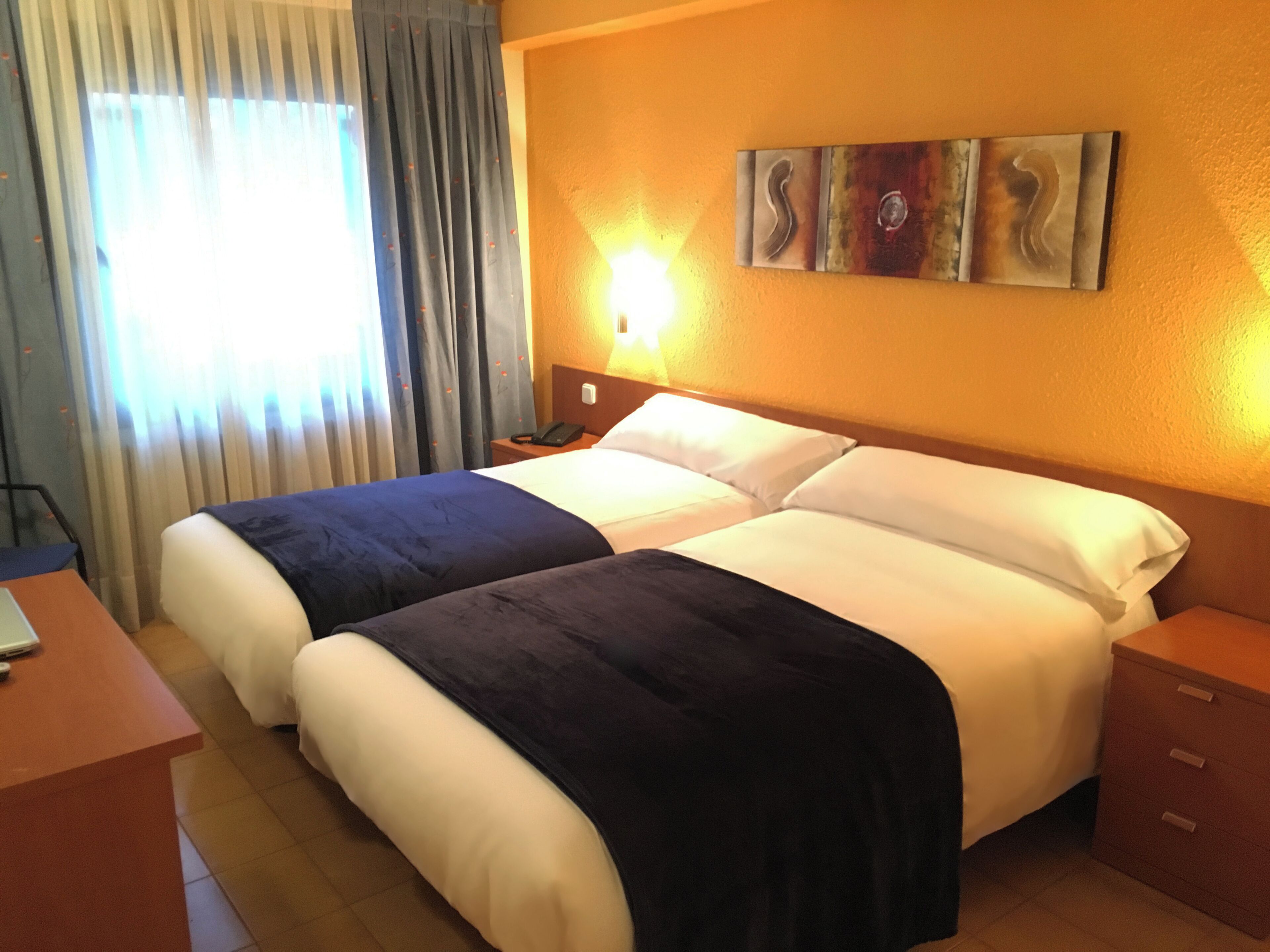 A hotel room with two beds, a desk, a nightstand, and a painting on the wall.