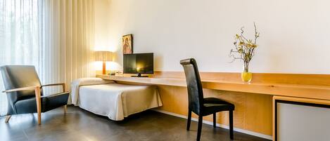 A hotel room with a bed, a desk with a computer, a chair, a lamp, and a vase with flowers.