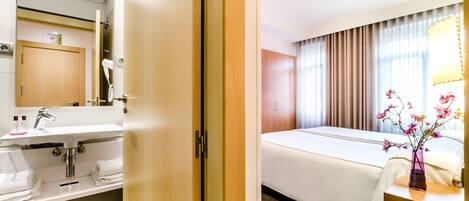 Une chambre d’hôtel avec une porte en bois, un lavabo blanc avec un miroir, un lit recouvert d’une courtepointe blanche et un vase de fleurs posé sur une table de chevet.