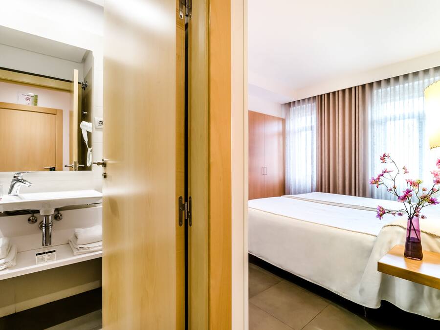 Habitación de hotel con puerta de madera, un lavabo blanco con espejo, una cama con cubrecama blanco y un florero con flores sobre una mesita de noche.