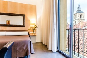 A hotel room with a bed, a bedside table with a lamp, a mirror, and a view of a building through a window. - Hospedería Pax (León)
