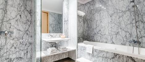 Un baño moderno con paredes de mármol, cabina de ducha con paredes de vidrio, tocador blanco y un banco.