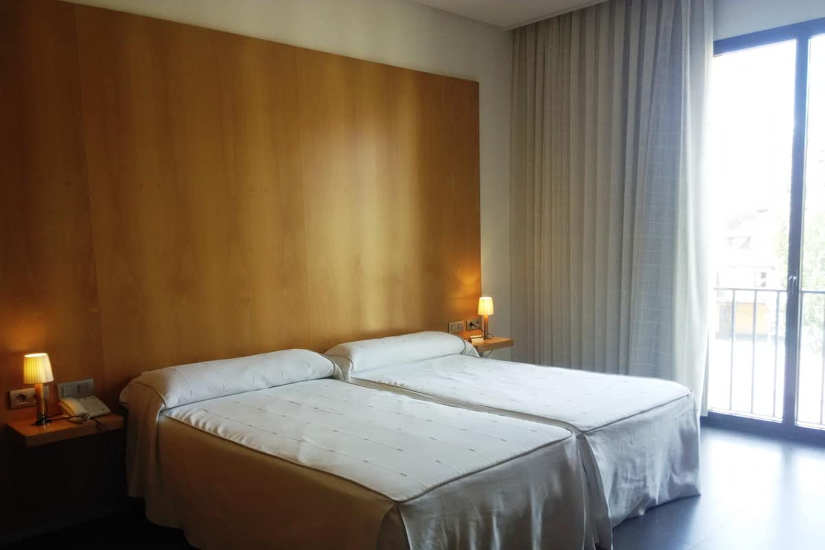 Habitación de hotel con dos camas, un cabecero de madera, una mesita de noche con lámpara y un ventanal con cortinas.