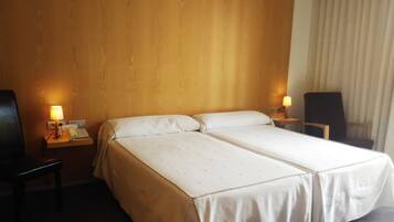 Habitación de hotel con dos camas individuales, cabecera de madera, lámparas de noche, una silla y un escritorio con teléfono.