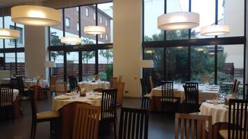 Een restaurant met meerdere tafels klaar voor diners, grote ramen met uitzicht op buiten, en hangende hanglampen.