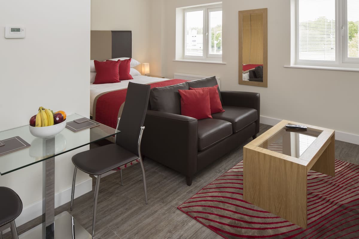 Moderni hotellihuone, jossa on lasinen ruokapöytä, punaisilla koristetyynyillä varustettu nahkasohva, puinen sohvapöytä ja suuri peili seinällä.