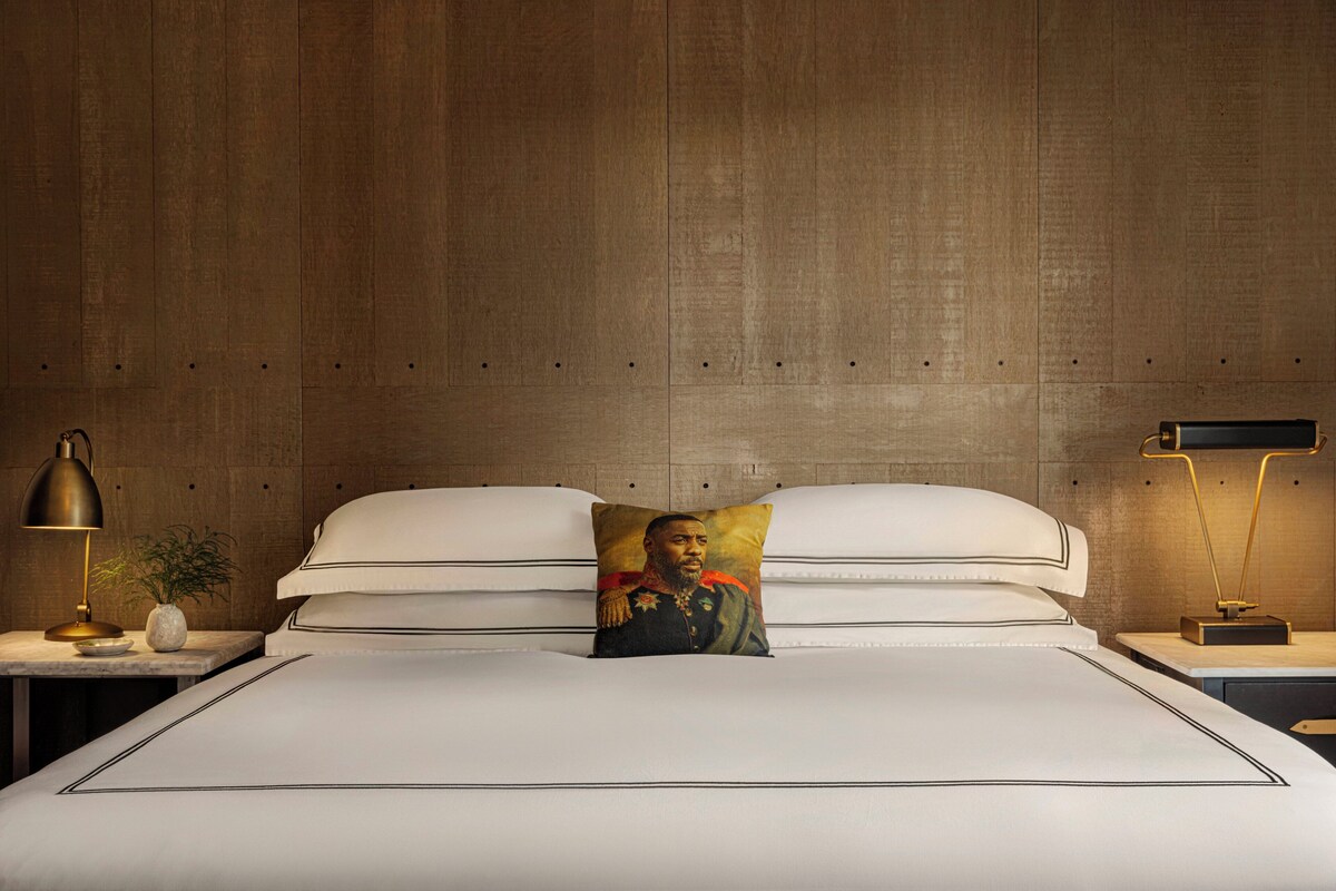 A neatly made bed with white linens, a decorative pillow, and a nightstand with a lamp and a small plant.