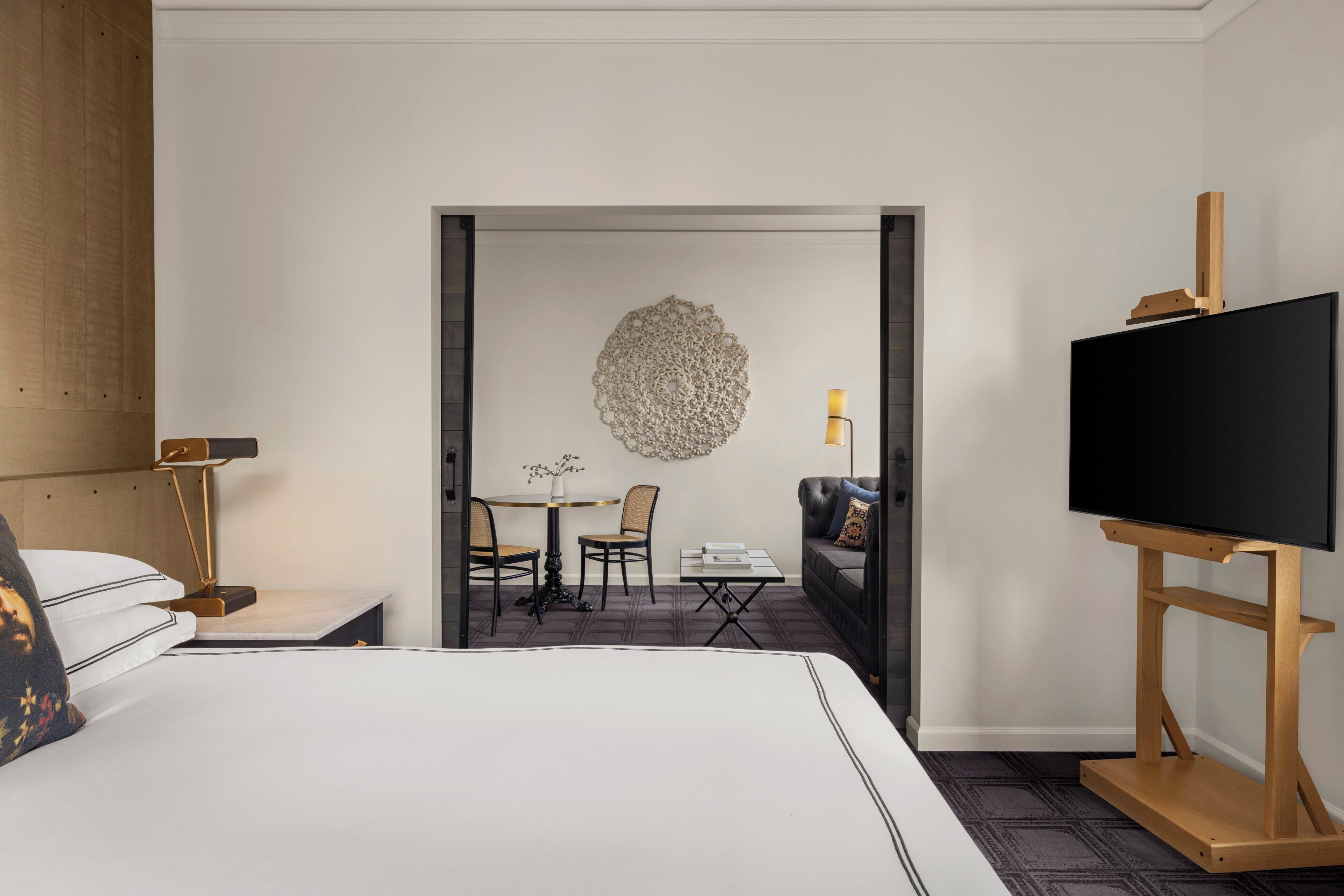 Habitación de hotel con una cama grande, un televisor sobre un soporte de madera, un escritorio con lámpara, un rincón de comedor con sillas y mesa, y un adorno en la pared.