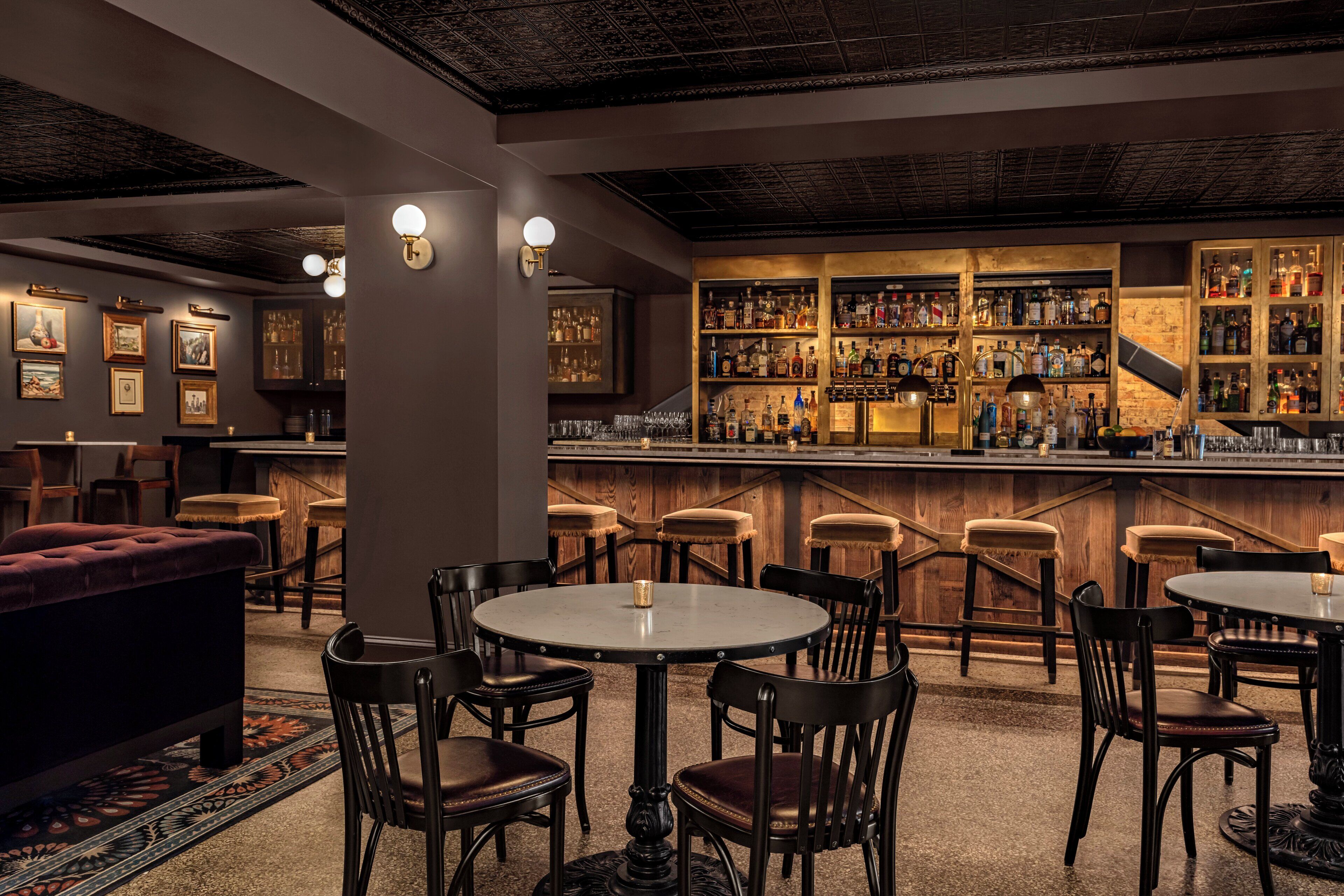Una zona de bar con mesas y sillas redondas, una barra con estantes llenos de botellas y cuadros en la pared.