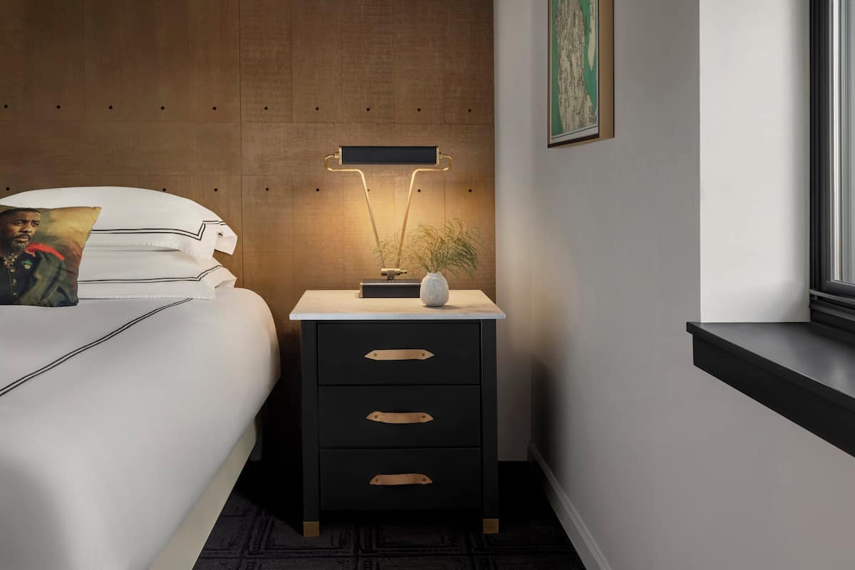 A neatly made bed with white linens and a pillow, a bedside table with a lamp, and a framed picture on the wall.