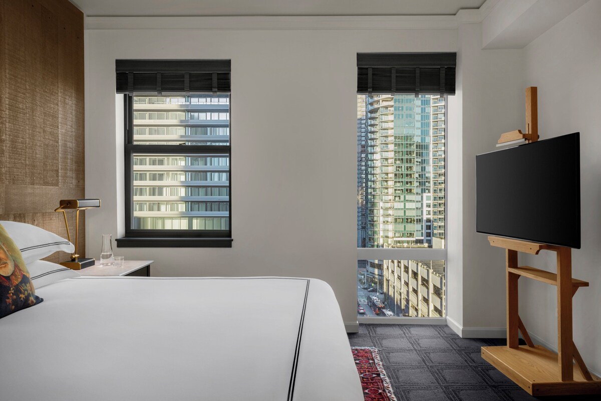 A modern hotel room with a large bed, a flat-screen TV on a wooden stand, a nightstand with a lamp, and a view of a high-rise building through a window.