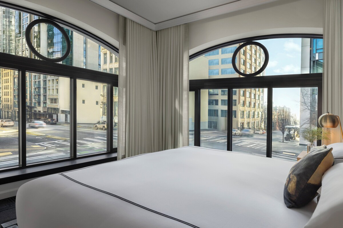 A modern hotel room with a large bed, a view of the city through floor-to-ceiling windows, and a circular window frame.