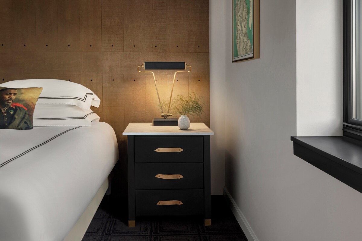 A neatly made bed with white linens and a pillow, a bedside table with a lamp, and a framed picture on the wall.