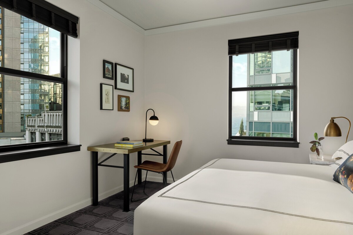 A hotel room with a bed, a desk with a chair, a lamp, and framed pictures on the wall.