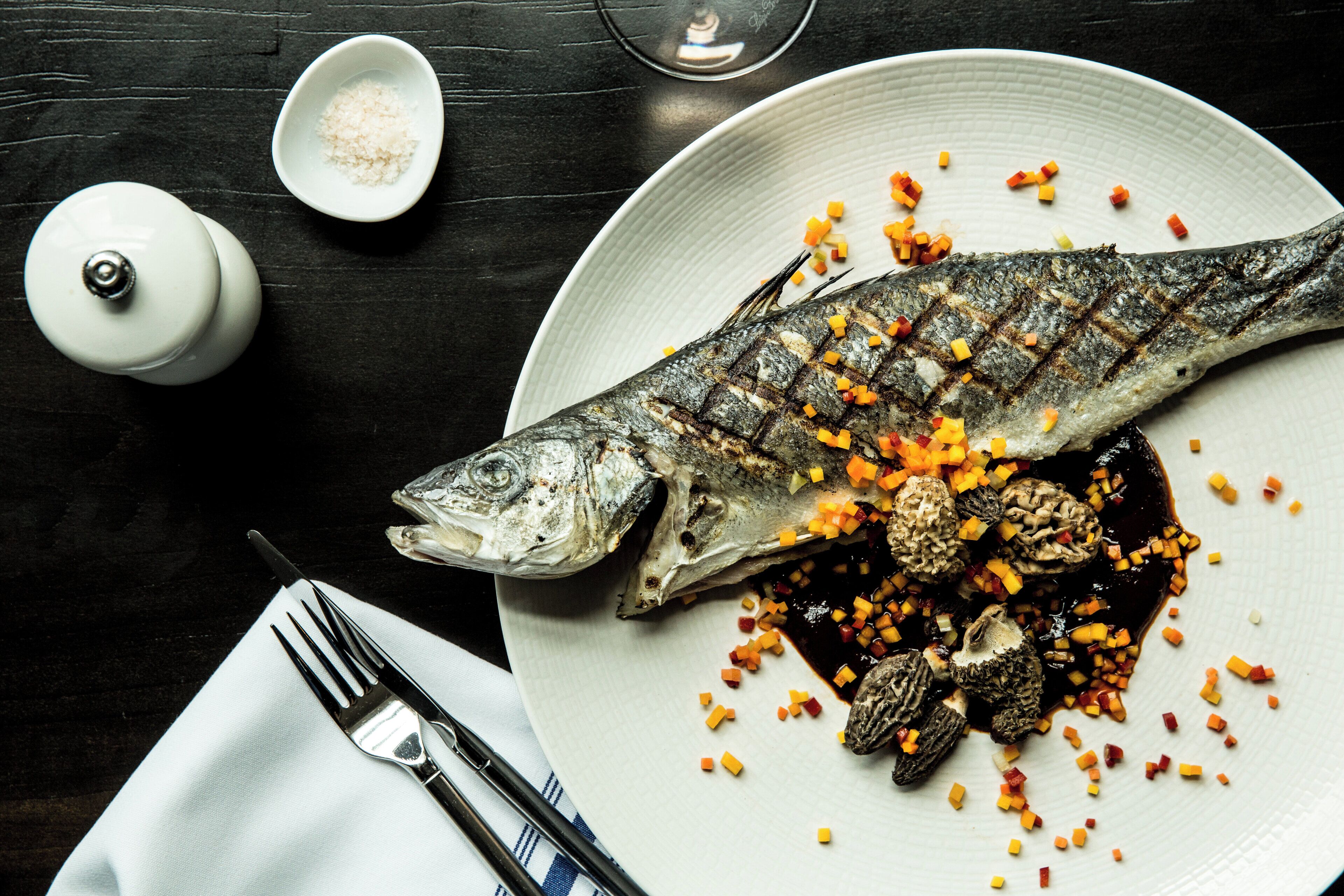 A plate with grilled fish, diced vegetables, and mushrooms on a dark wooden table.