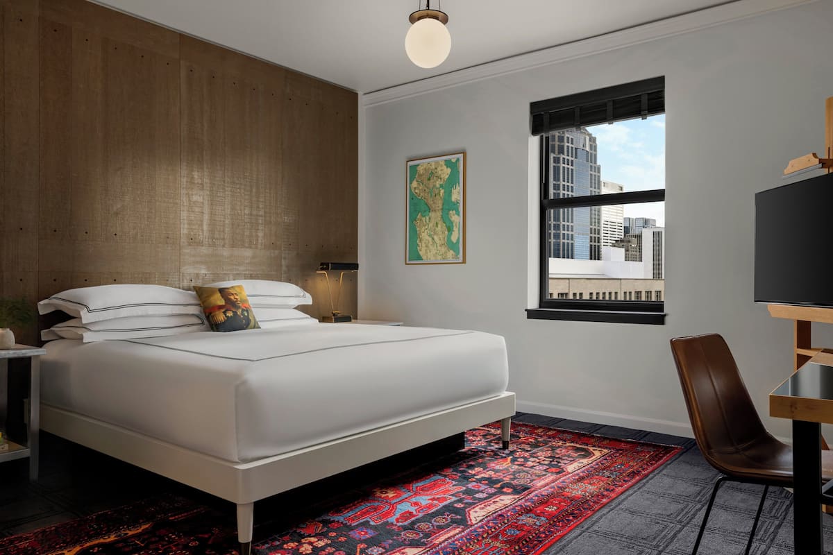 A modern bedroom with a large bed, a wooden accent wall, a city view through a window, and a red patterned rug.