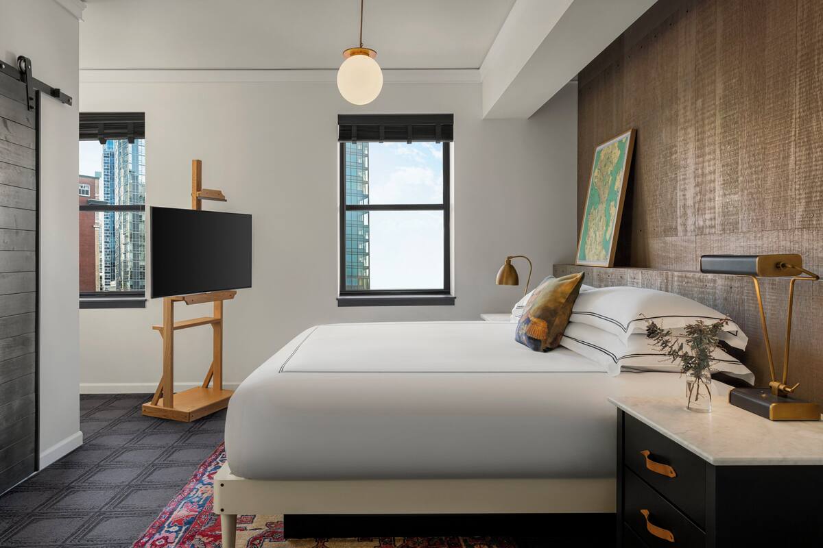 A modern bedroom with a large bed, a desk with a lamp, a map on the wall, and a view of buildings through the window.